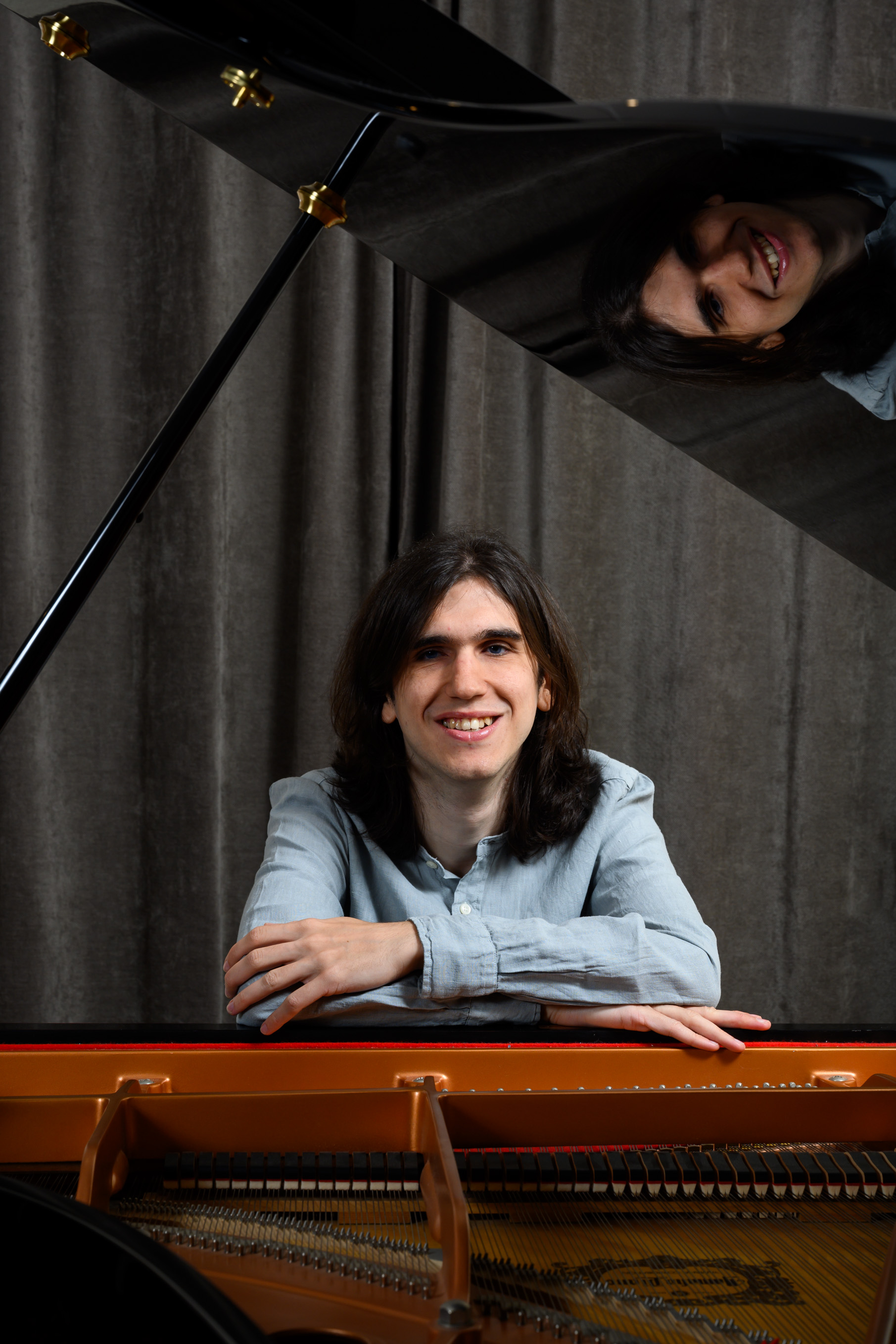 Guillem León Vivas leaning on the open lid of a grand piano, smiling, his face reflected in the lacquer above him.